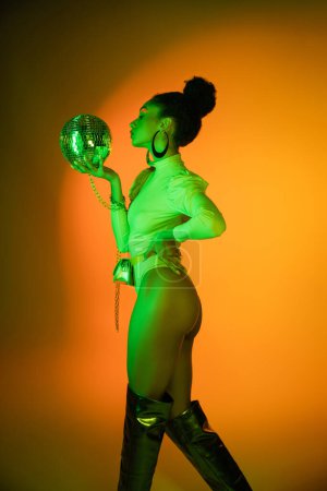 Foto de Vista lateral de la sexy mujer afroamericana en traje de neón mirando la bola disco sobre fondo naranja - Imagen libre de derechos