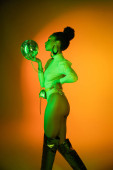 Side view of sexy african american woman in neon bodysuit looking at disco ball on orange background  magic mug #648332908