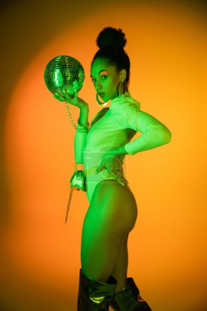 Trendy african american model in neon bodysuit and knee boots holding disco ball and looking at camera on orange background 