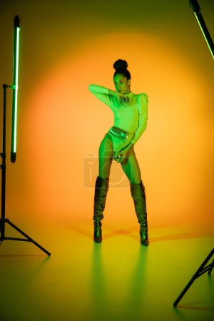 African american model in neon bodysuit and makeup holding disco balls near fluorescent lamps on orange background 