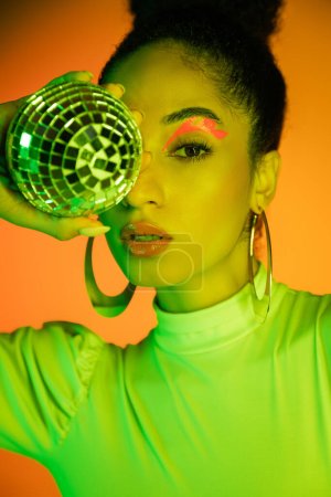 Portrait of trendy african american model with neon visage holding disco ball on orange background 