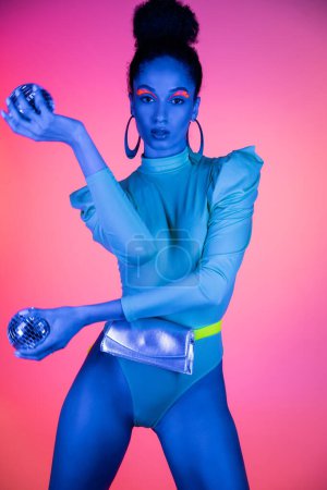 Fashionable african american model with neon makeup and bodysuit holding disco balls on pink background