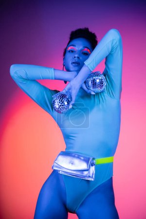 Modèle afro-américain à la mode avec eye-liner néon posant avec des boules de disco sur fond rose et violet