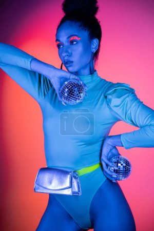 Trendy african american woman in neon bodysuit holding disco balls on pink background