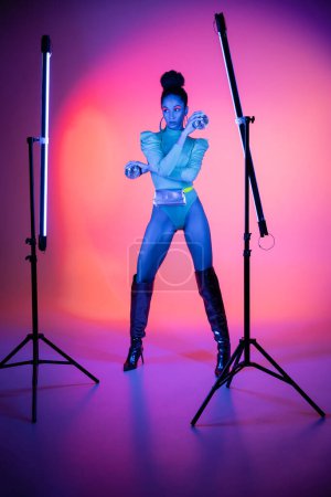 Modisches afrikanisch-amerikanisches Model mit Discokugeln in der Nähe von Neonlampen auf lila Hintergrund