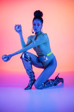 Trendy african american woman in bodysuit holding disco balls on pink and purple background with neon light 
