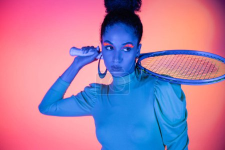 Fashionable african american woman with neon eyeliner holding tennis racket on pink background
