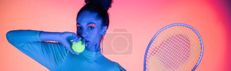 Pretty african american model with neon visage holding ball and tennis racket on pink background, banner 