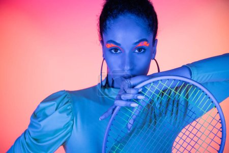 Retrato de mujer afroamericana de moda con rostro de neón sosteniendo raqueta de tenis sobre fondo rosa claro