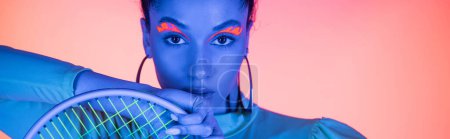 Trendy african american woman with neon eyeliner holding tennis racket on light pink background, banner 