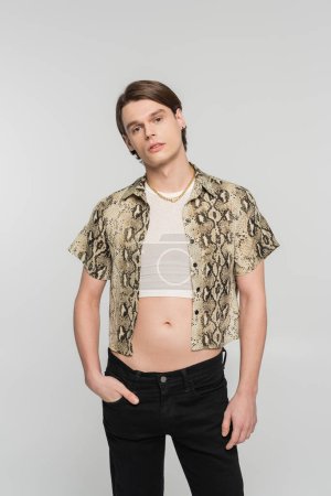 stylish bigender person in snakeskin print blouse posing with hand in pocket of black pants isolated on grey
