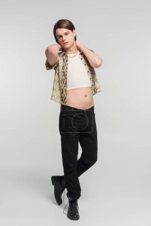 full length of bigender model in black pants and snakeskin print blouse posing with hands behind neck on grey background