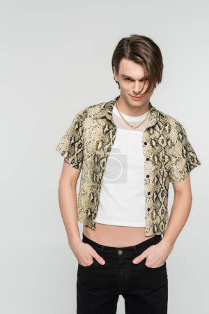 young and trendy bigender model in animal print blouse posing with hands in pockets of black pants isolated on grey