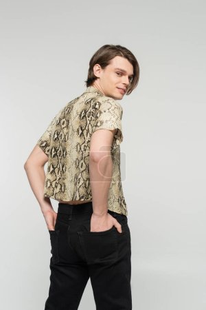 trendy pangender person in animal print blouse holding hands in back pockets and smiling at camera isolated on grey