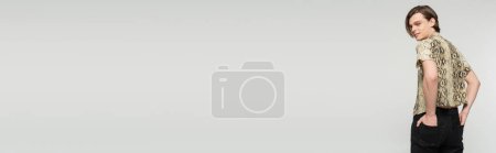 Photo for Happy nonbinary person holding hands in back pockets and smiling at camera isolated on grey, banner - Royalty Free Image