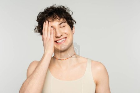 cheerful nonbinary model in tank top and beads covering face with hand and smiling with closed eyes isolated on grey