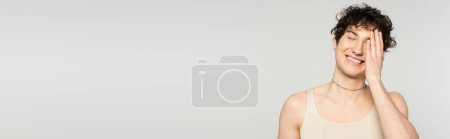 cheerful pansexual model in tank top covering face with hand and smiling with closed eyes isolated on grey, banner