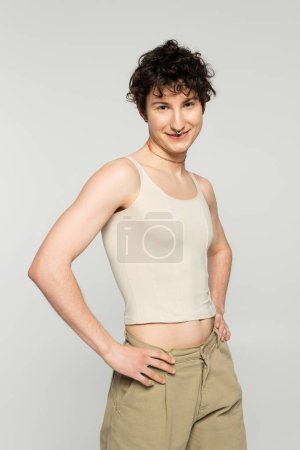 trendiges und glückliches Pangender-Model, das mit den Händen auf den Hüften steht und isoliert auf grau in die Kamera lächelt