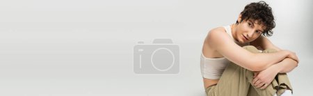 Photo for Stylish nonbinary model sitting and hugging knees while looking away isolated on grey, banner - Royalty Free Image