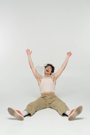 excited nonbinary person in beige pants laughing while sitting with raised hands on grey background