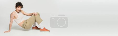 in voller Länge trendiges Pangender-Modell in beiger Hose und Gummischuhen, das auf grauem Hintergrund in die Kamera schaut, Banner