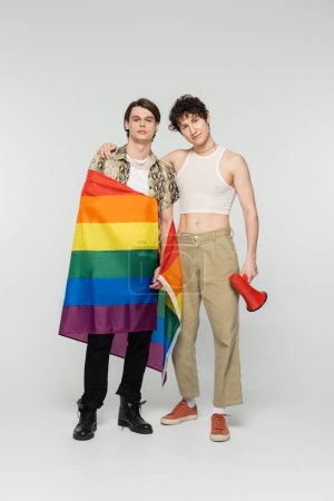 full length of young nonbinary couple with lgbt flag and megaphone looking at camera on grey background