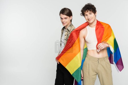 glückliches und stylisches Pangender-Paar mit Regenbogenfahne, das isoliert auf grau in die Kamera lächelt