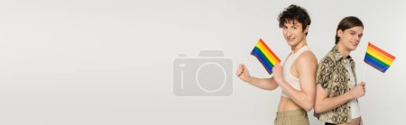 Téléchargez les photos : Heureux couple non binaire tenant petits drapeaux lgbt et souriant à la caméra isolé sur gris, bannière - en image libre de droit