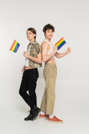 Foto de Longitud completa de elegantes modelos pansexuales posando con pequeñas banderas lgbt sobre fondo gris - Imagen libre de derechos