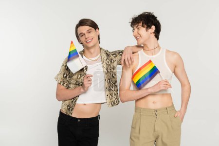 cheerful and stylish bigender models posing with small lgbt flags isolated on grey