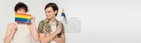 cheerful pansexual person looking at partner obscuring face with small lgbt flag isolated on grey, banner