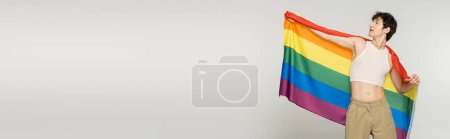 Téléchargez les photos : Brunette modèle non binaire en haut de la culture posant avec drapeau lgbt isolé sur gris, bannière - en image libre de droit