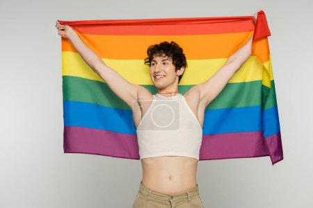 joyful bigender model in crop top holding lgbt flag in raised hands and looking away isolated on grey