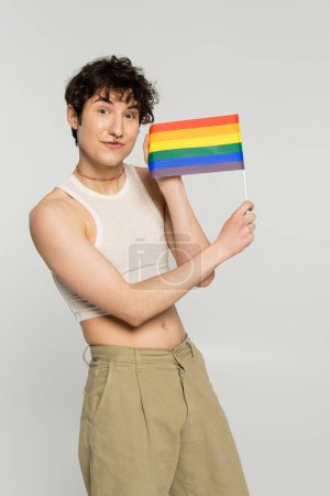 Téléchargez les photos : Gai brunette modèle pansexuel dans le haut de la culture montrant petit drapeau lgbt isolé sur gris - en image libre de droit