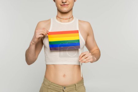 partial view of young pangender model in crop top holding small lgbt flag isolated on grey