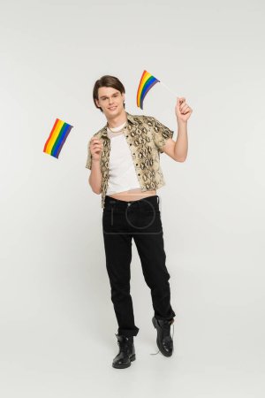 full length of positive pansexual model in black pants and snakeskin print blouse holding small lgbt flags on grey background