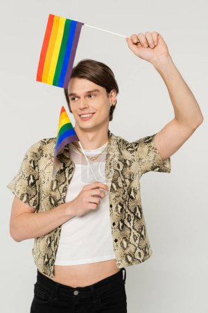 Foto de Modelo pansexual despreocupado en elegante blusa posando con pequeñas banderas lgbt aisladas en gris - Imagen libre de derechos