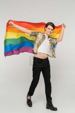 Foto de Longitud completa de la persona no binaria elegante y despreocupada caminando con la bandera del arco iris sobre fondo gris - Imagen libre de derechos
