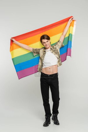 full length of carefree and trendy bigender person holding rainbow flag in raised hands on grey background