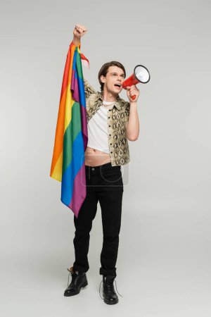 full length of bigender person in animal print blouse holding lgbt flag and shouting in megaphone on grey background