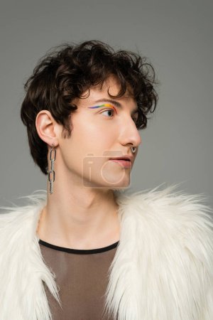 Porträt einer stilvollen pansexuellen Person mit silbernem Ohrring und Nasenpiercing, die weiße Kunstpelzjacke isoliert auf grau trägt