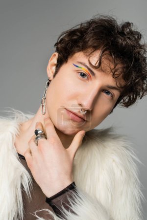portrait of pansexual model with wavy brunette hair and rainbow eye liner looking at camera isolated on grey