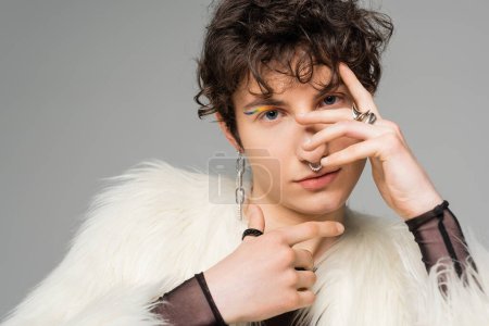 brunette pansexual person in white faux fur jacket and silver accessories obscuring face with hand isolated on grey