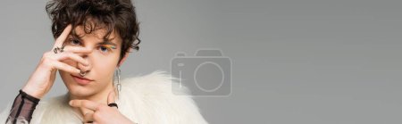 portrait of stylish bigender person in white faux fur jacket covering face with hand isolated on grey, banner