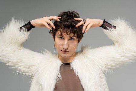 brunette nonbinary model in silver accessories and white faux fur jacket posing with hands near head isolated on grey