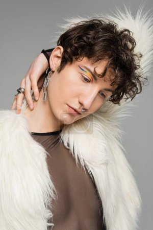 Photo for Portrait of fashionable bigender person with rainbow eye liner posing in white faux fur jacket isolated on grey - Royalty Free Image
