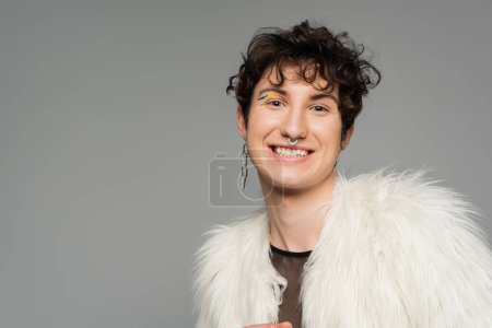 overjoyed pansexual model in silver accessories and white faux fur jacket smiling at camera isolated on grey