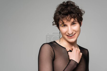 Photo for Happy bigender person in black transparent top and silver accessories looking at camera isolated on grey - Royalty Free Image