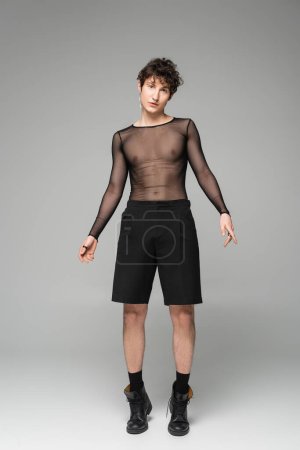 longitud completa de la persona no binaria en top transparente y pantalones cortos negros sobre fondo gris