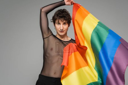 Photo for Brunette bigender person in black transparent top holding rainbow flag isolated on grey - Royalty Free Image